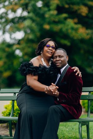 A couple in formal attire shares an intimate moment on a park bench surrounded by greenery.