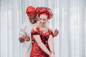 A newly married couple in matching red traditional African-inspired attire dance and laugh together against a white curtain.