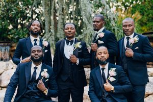 Wedding party in formal black suits and boutonnieres poses together outdoors on garden pathway.