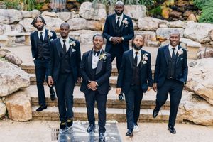 Wedding groomsmen in crisp black suits walking in line against a natural stone backdrop outdoors.