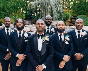 Group of groomsmen in black tuxedos with white boutonnieres posing together outdoors.