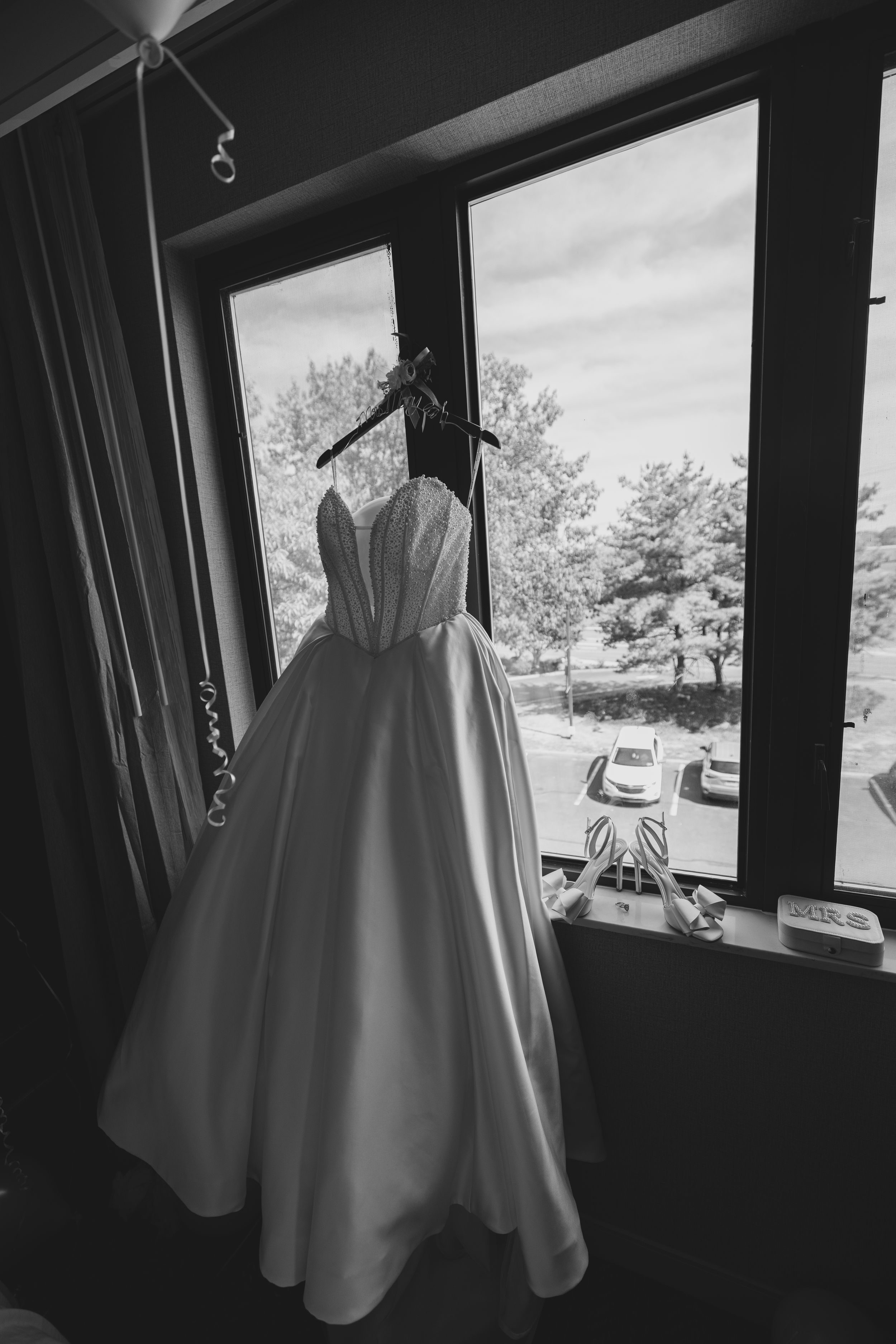 A white wedding dress hangs in front of a window, creating a series of silhouettes in black and white photography.