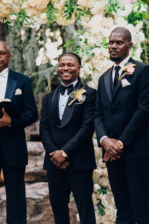 Groomsmen in classic black tuxedos stand together outdoors during a wedding celebration.