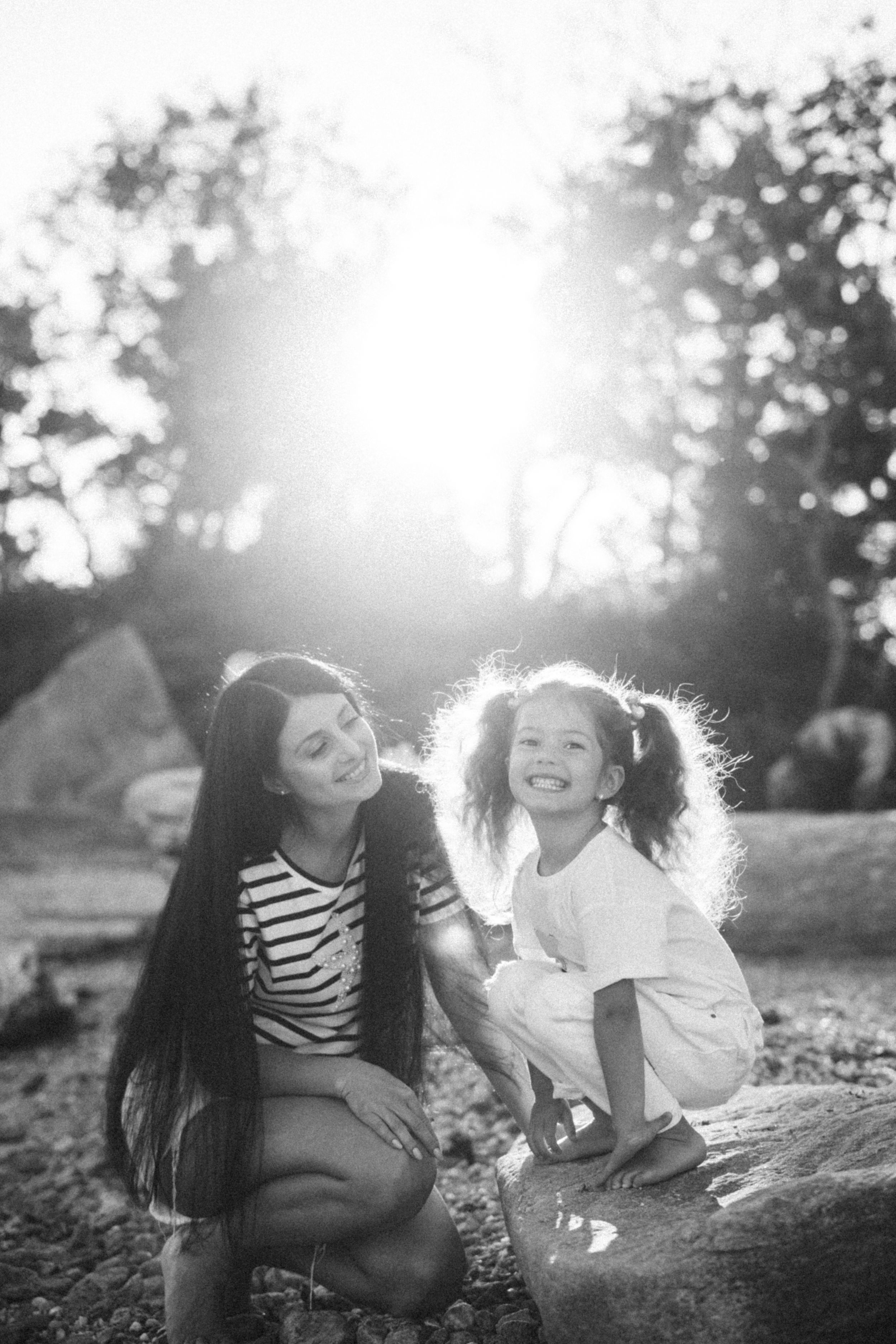 Black and white photo of two people enjoying a candid moment outdoors with sunlight creating a backlit glow through trees.