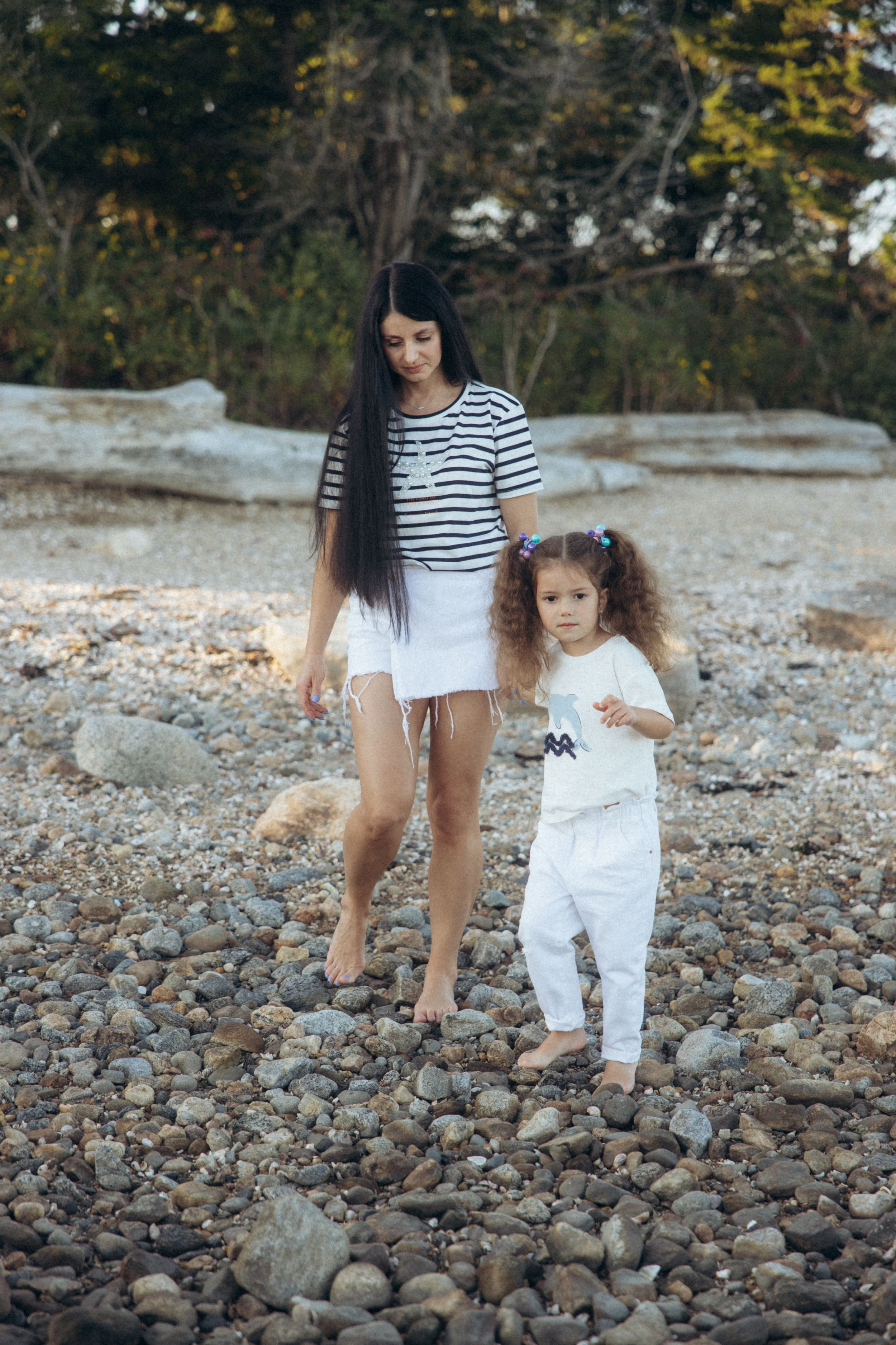 Two people in casual summer outfits walking together on a rocky beach surrounded by trees at sunset.