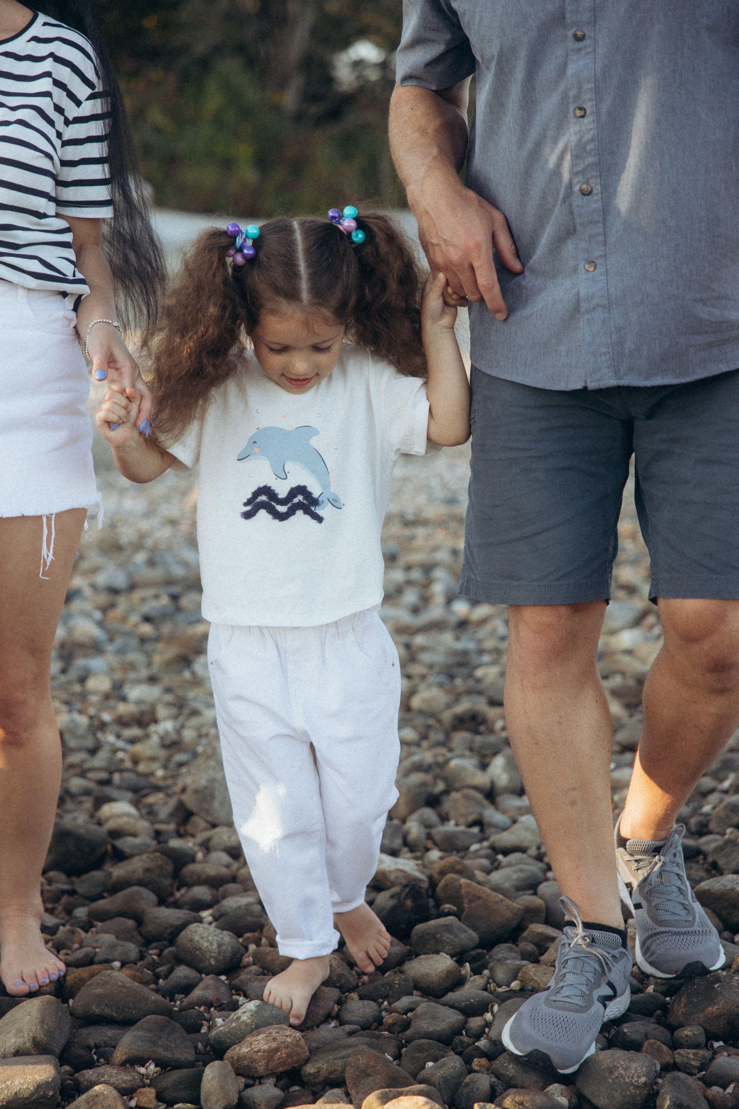 Child in white outfit with bear graphic walking on rocky beach while holding hands with adults viewing from waist down.