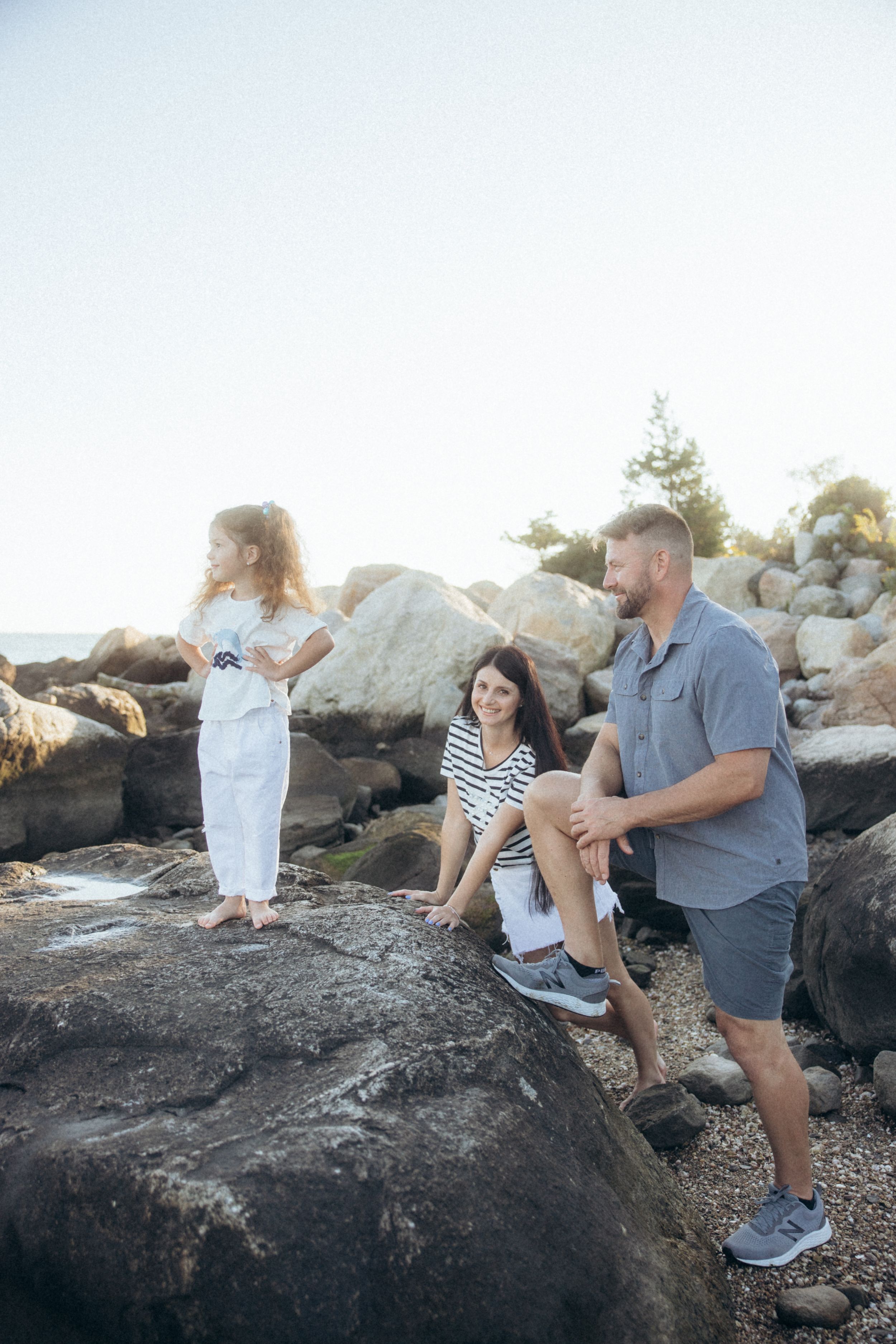A family explores rocky coastal terrain at sunset, dressed casually and standing on large boulders near the ocean.
