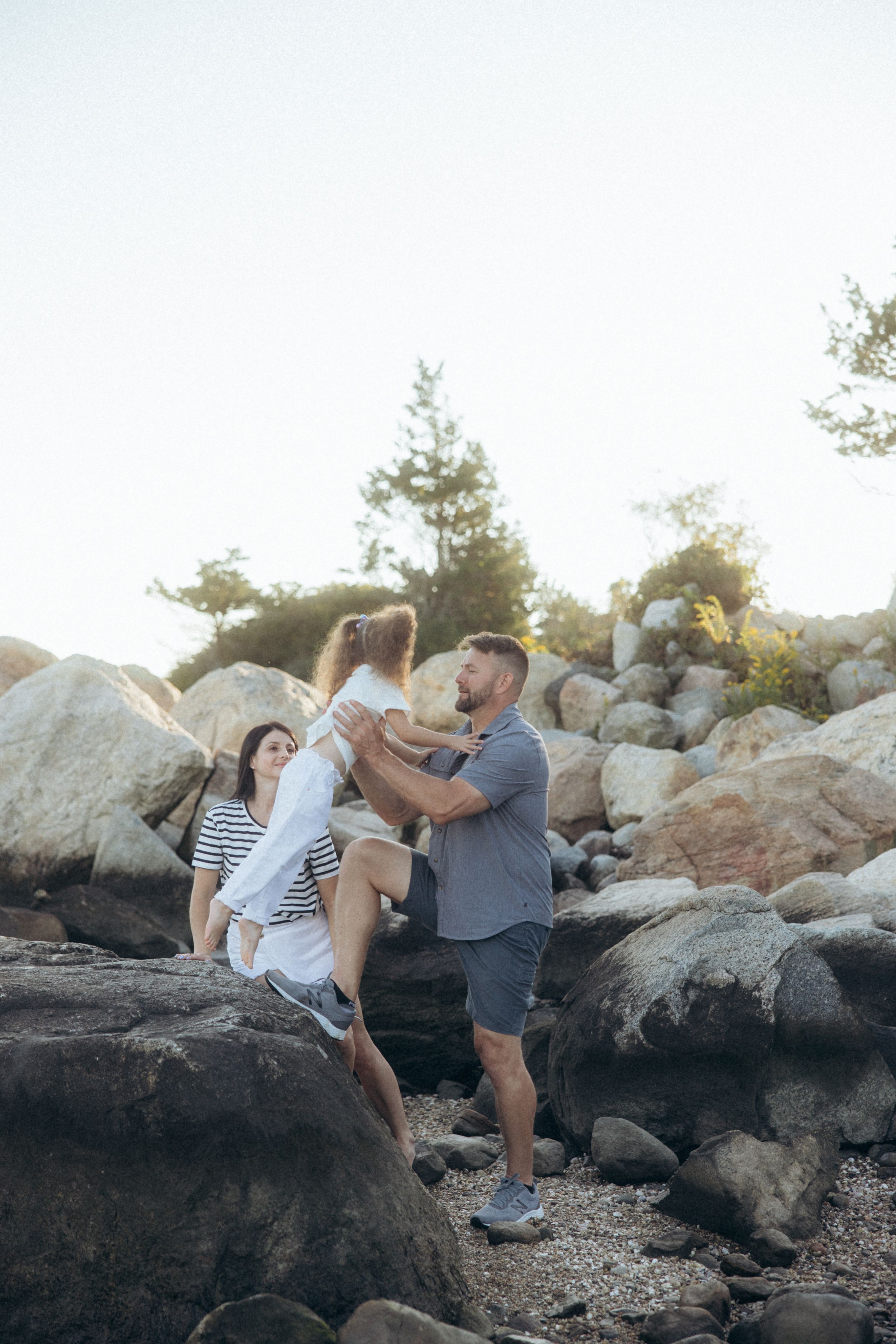 A family enjoys quality time together while climbing on large rocky boulders by the water's edge on a sunny day.