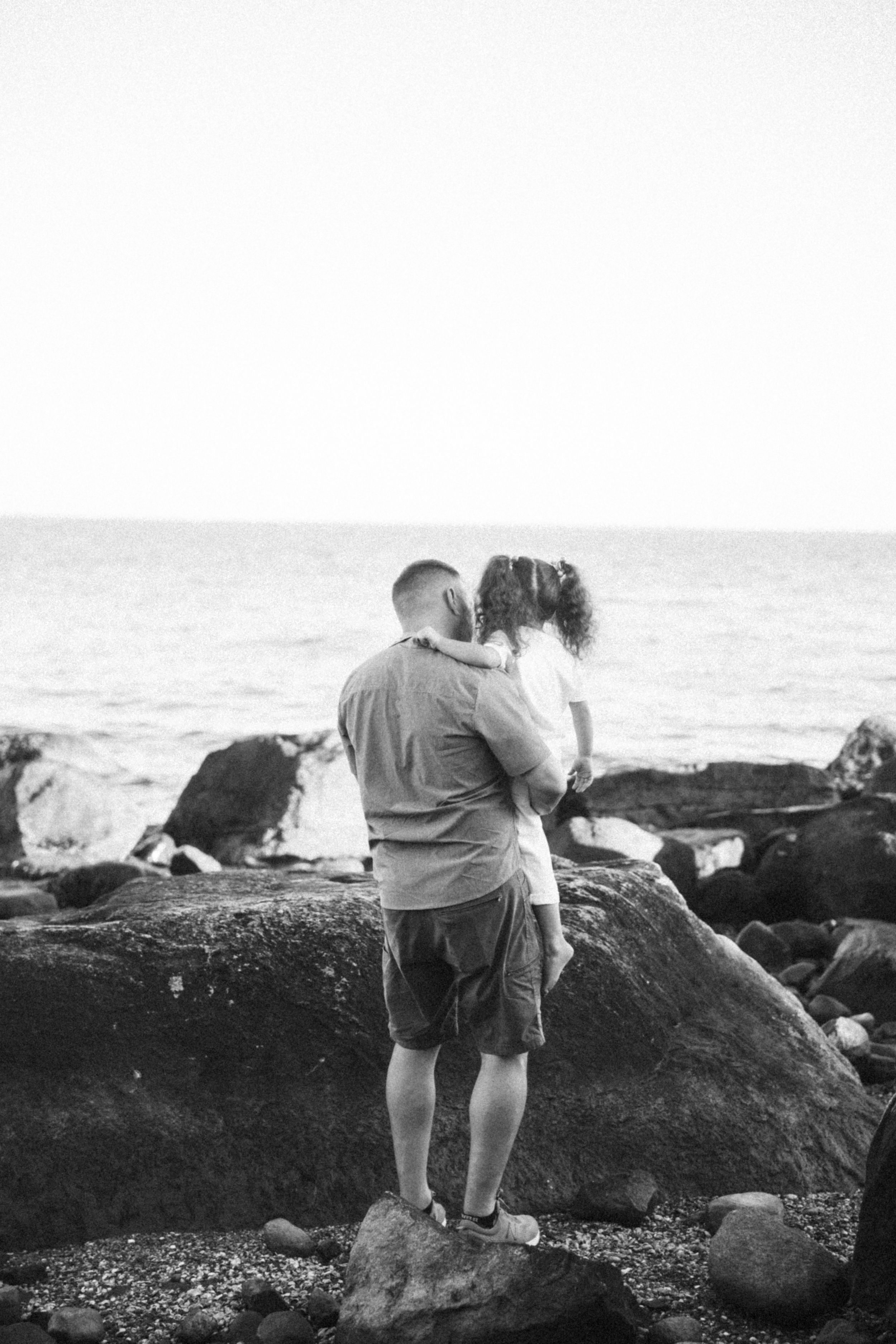 A loving moment shared between two people standing on coastal rocks overlooking the ocean at sunset.