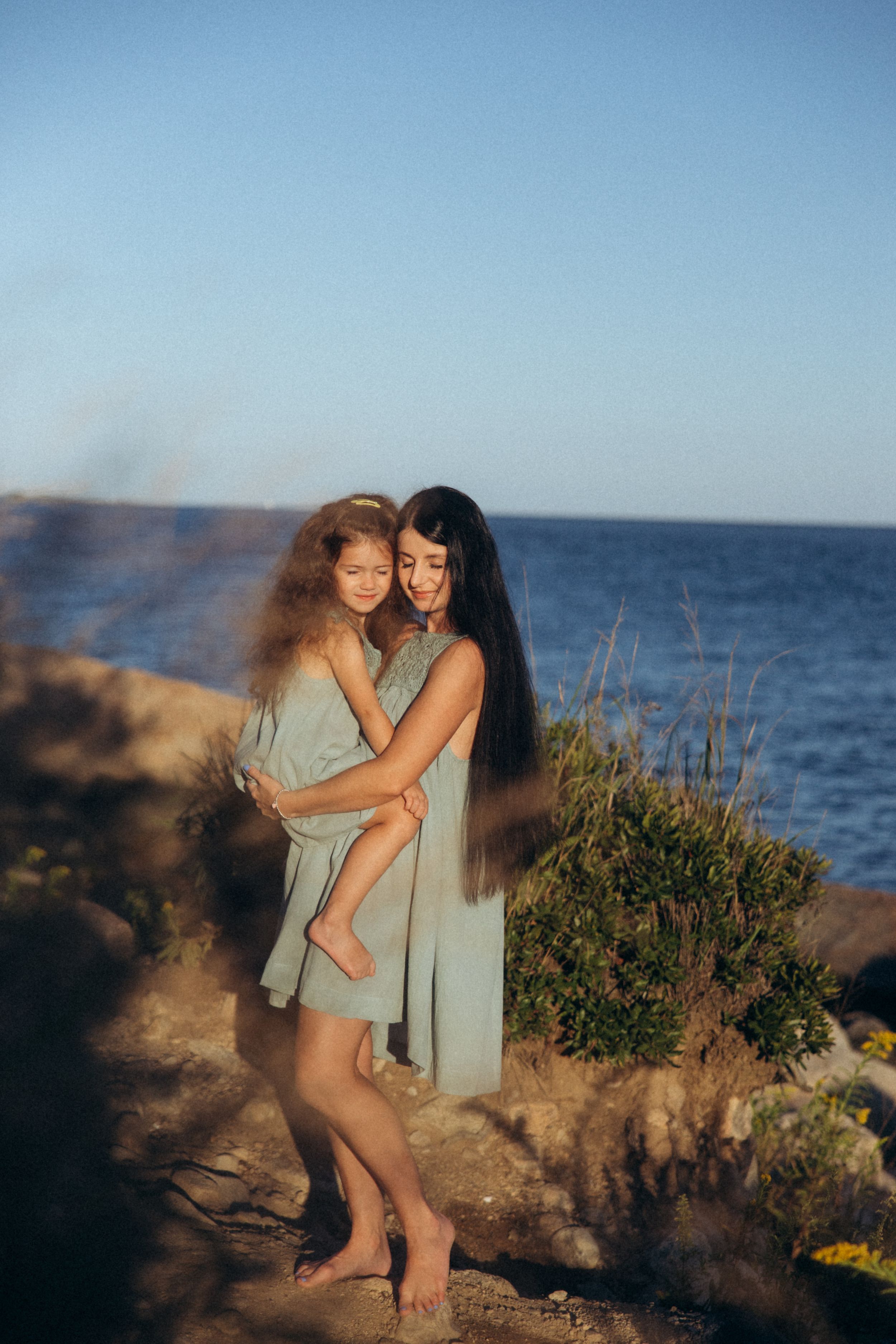 A tender moment shared between two people standing on rocky coastal cliffs overlooking the ocean at sunset.