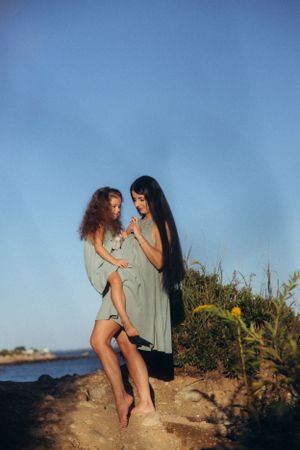 Two women in flowy dresses share an intimate moment on a coastal cliff against a bright blue sky.