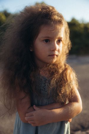 A young child with flowing curly hair wearing a gray dress stands in golden sunlight during a dreamy outdoor photoshoot.