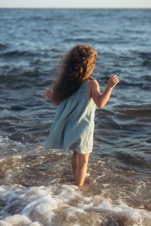 A small child in a light dress plays in ocean waves during golden hour.