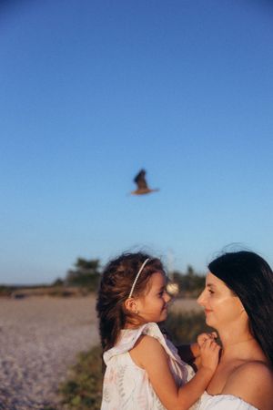 Two people share a tender moment on a beach at sunset while a bird soars overhead against the blue sky.