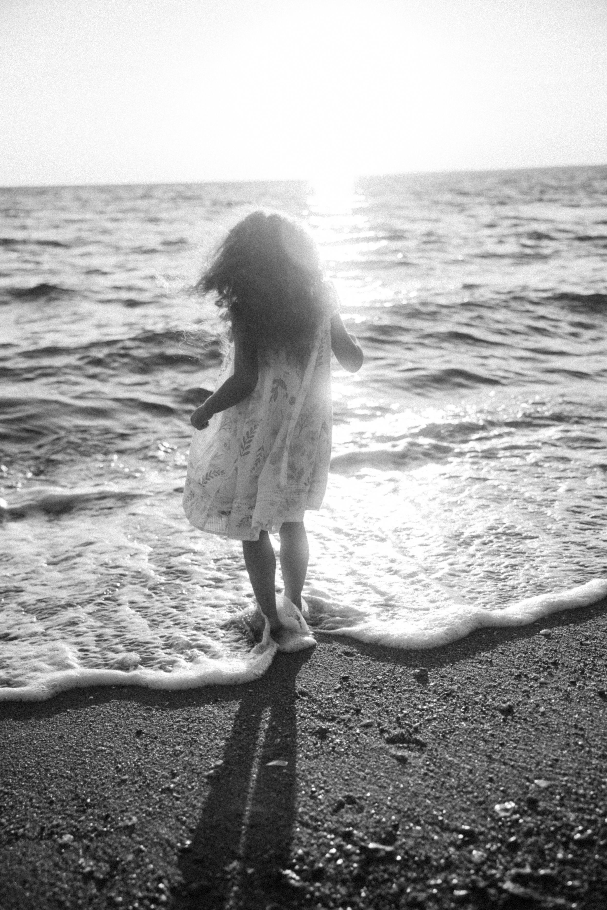 Silhouette figure in white dress stands at ocean's edge during sunset, creating long shadow on sandy beach.
