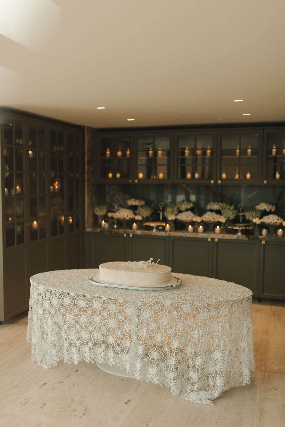Elegant wedding cake display with white lace tablecloth in a dimly lit reception venue with dark walls and ambient lighting.