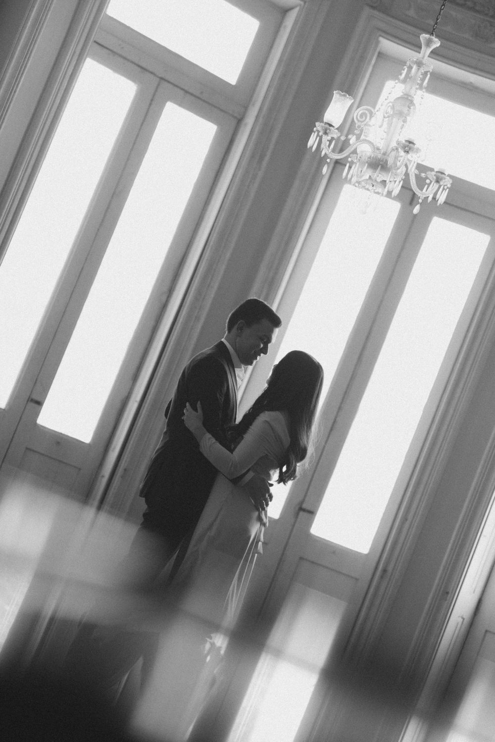 A romantic silhouette of a couple embracing near tall windows with a crystal chandelier in an elegant room.