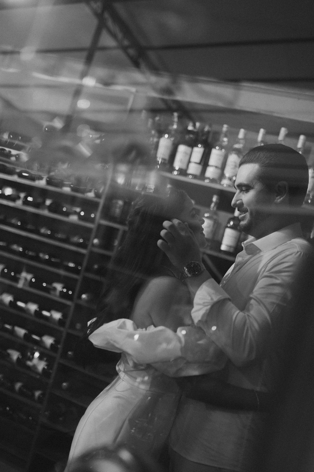 Black and white photo of a couple embracing in a wine cellar surrounded by bottle-filled shelves.