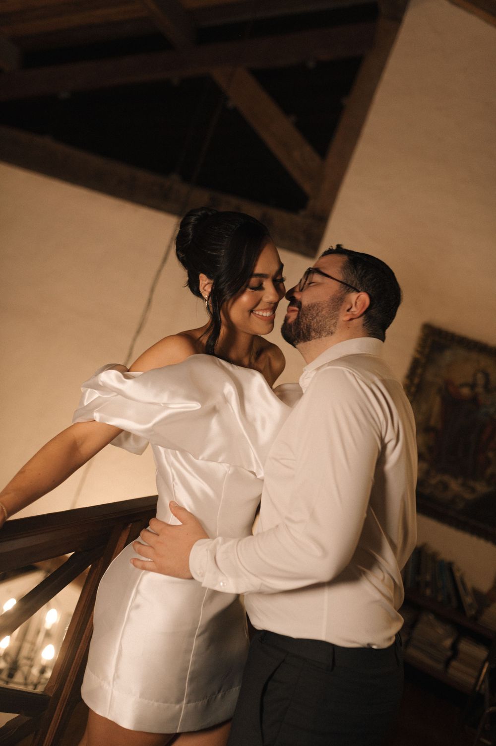Couple sharing a romantic embrace on a staircase, woman in white off-shoulder mini dress, man in white shirt.