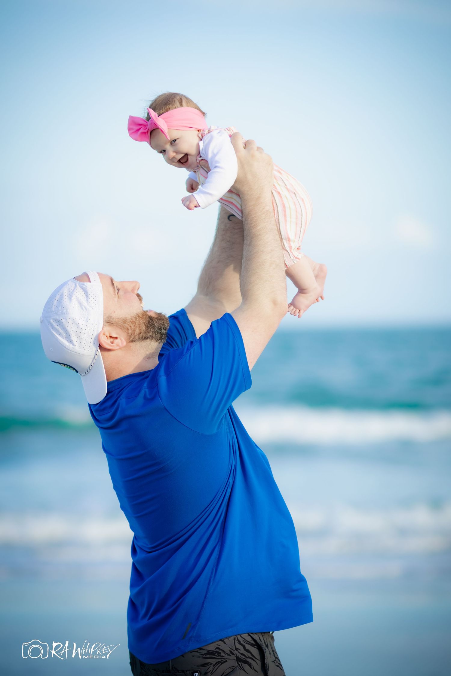 A heartwarming moment at the beach as someone lifts a baby wearing a pink bow against a blue ocean backdrop.