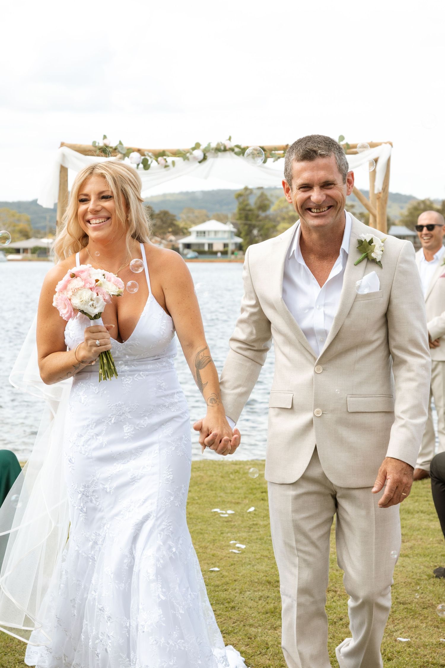 A newlywed couple walks hand in hand on a waterfront lawn with floral wedding arch and water views in the background.