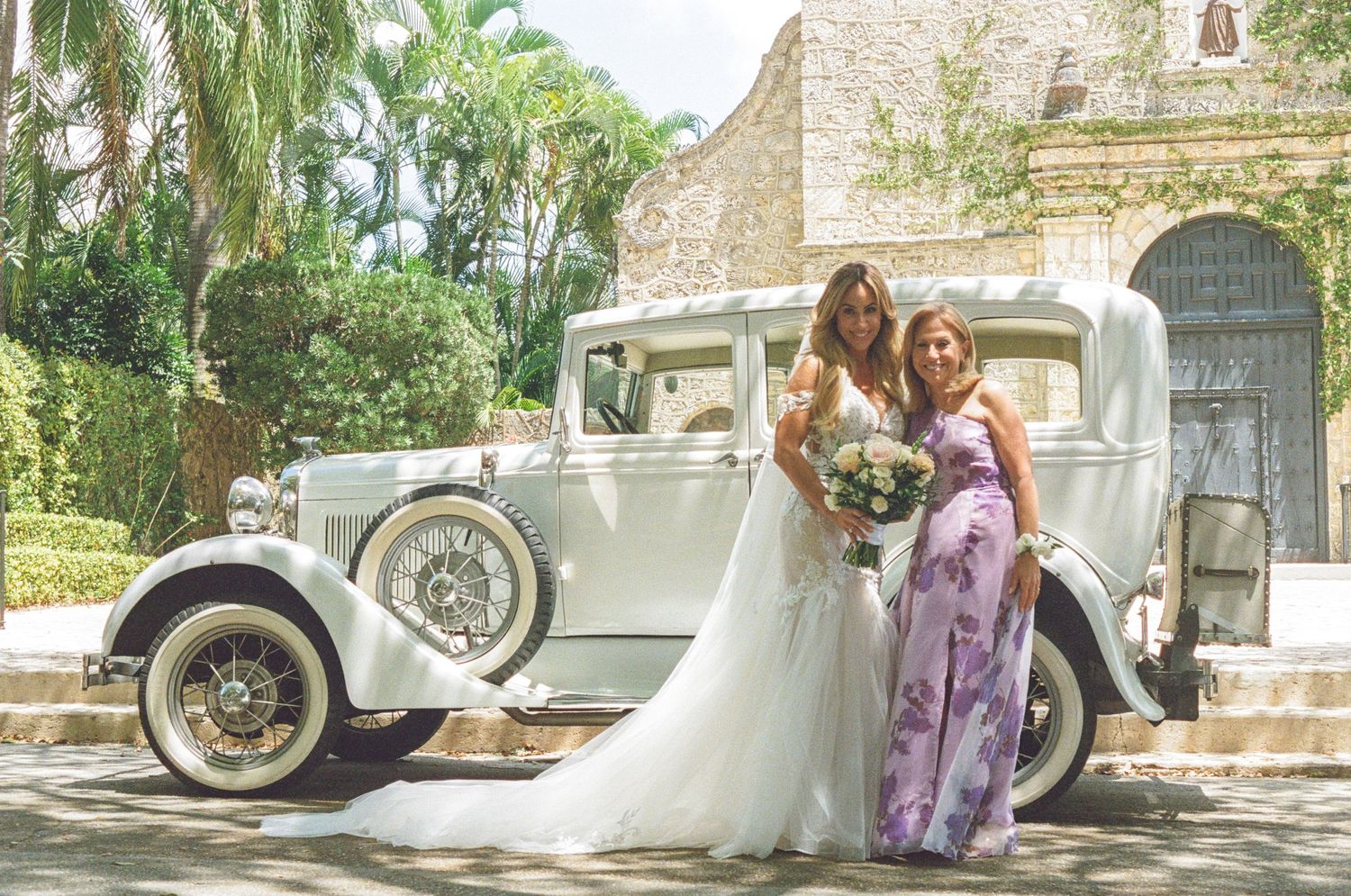 A vintage cream-colored luxury car parked in front of a stone church with palm trees and floral decorations.