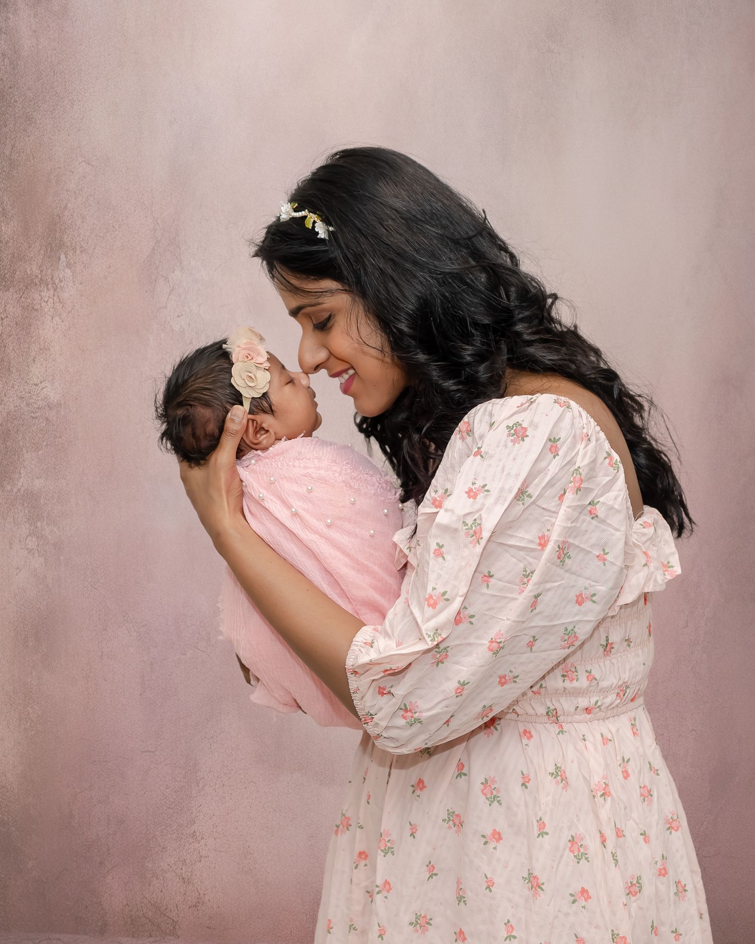 A woman in a floral dress smiles lovingly while holding a sleeping baby swaddled in pink against a pink background.