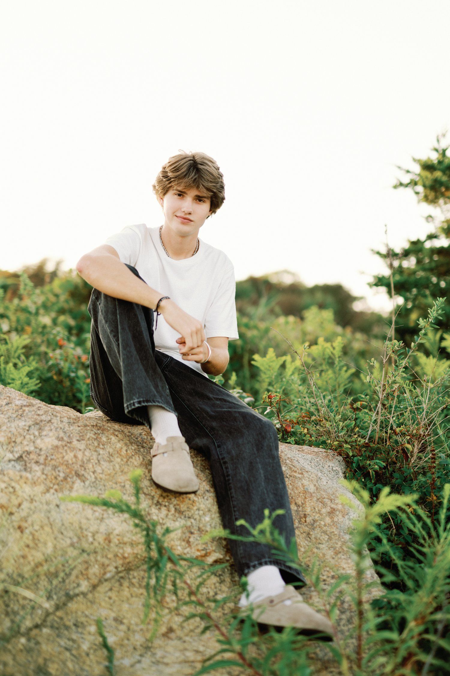 Someone sits on grass at sunset looking at phone while surrounded by lush green foliage in soft sunlight.