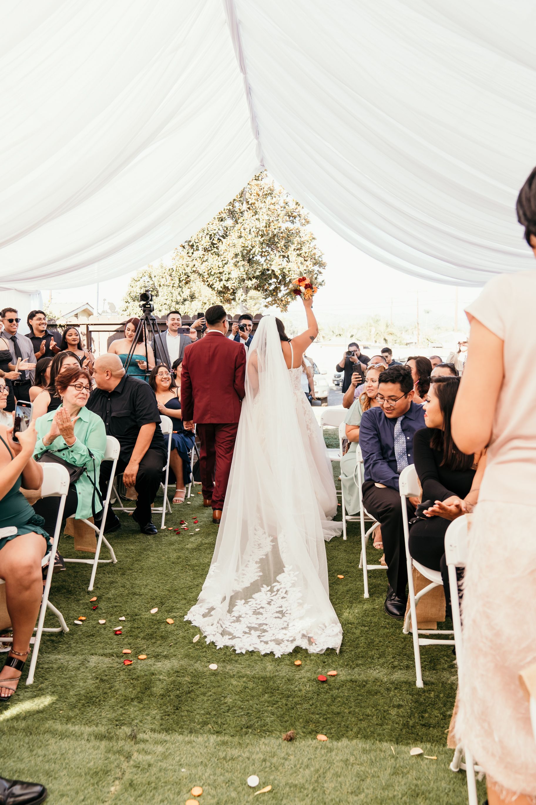 Wedding ceremony under a white tent with guests seated on folding chairs as couple walks down the aisle.