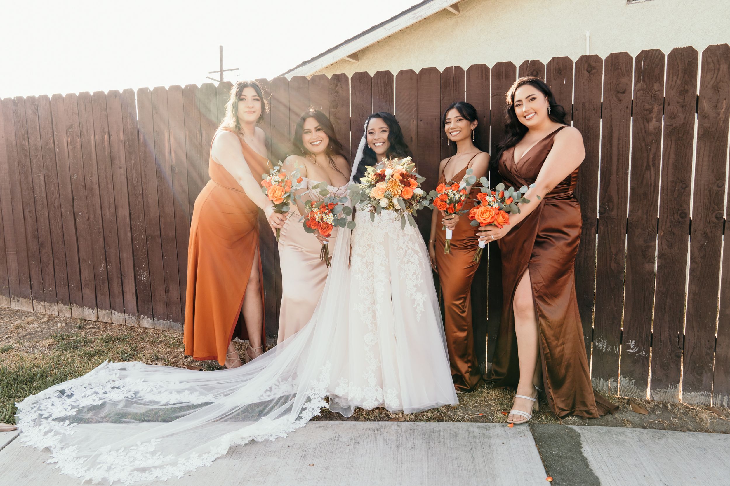 Wedding party in rust-colored dresses poses with bride in flowing white gown against wooden fence.