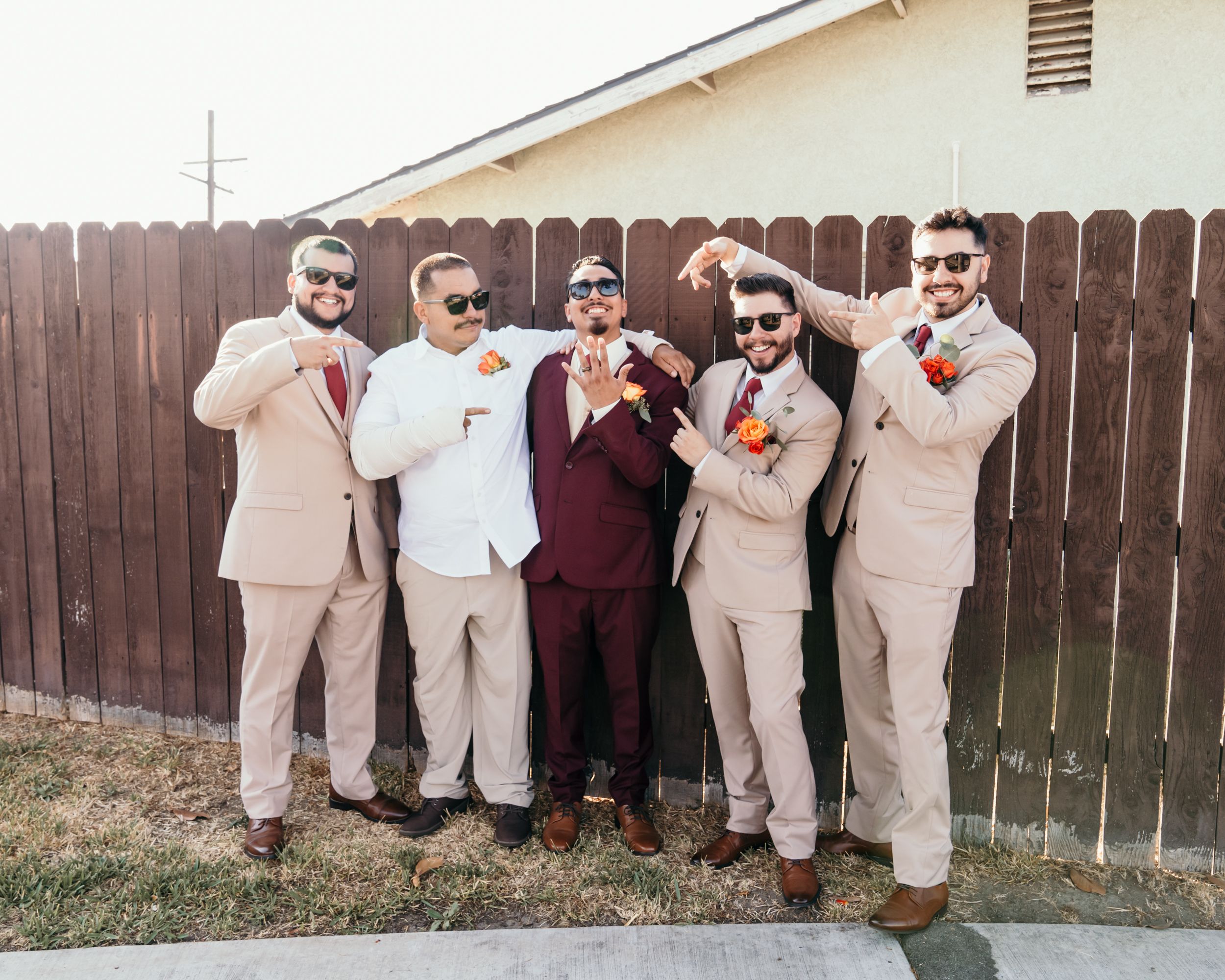 Wedding party in burgundy and tan suits posing playfully against a wooden fence.