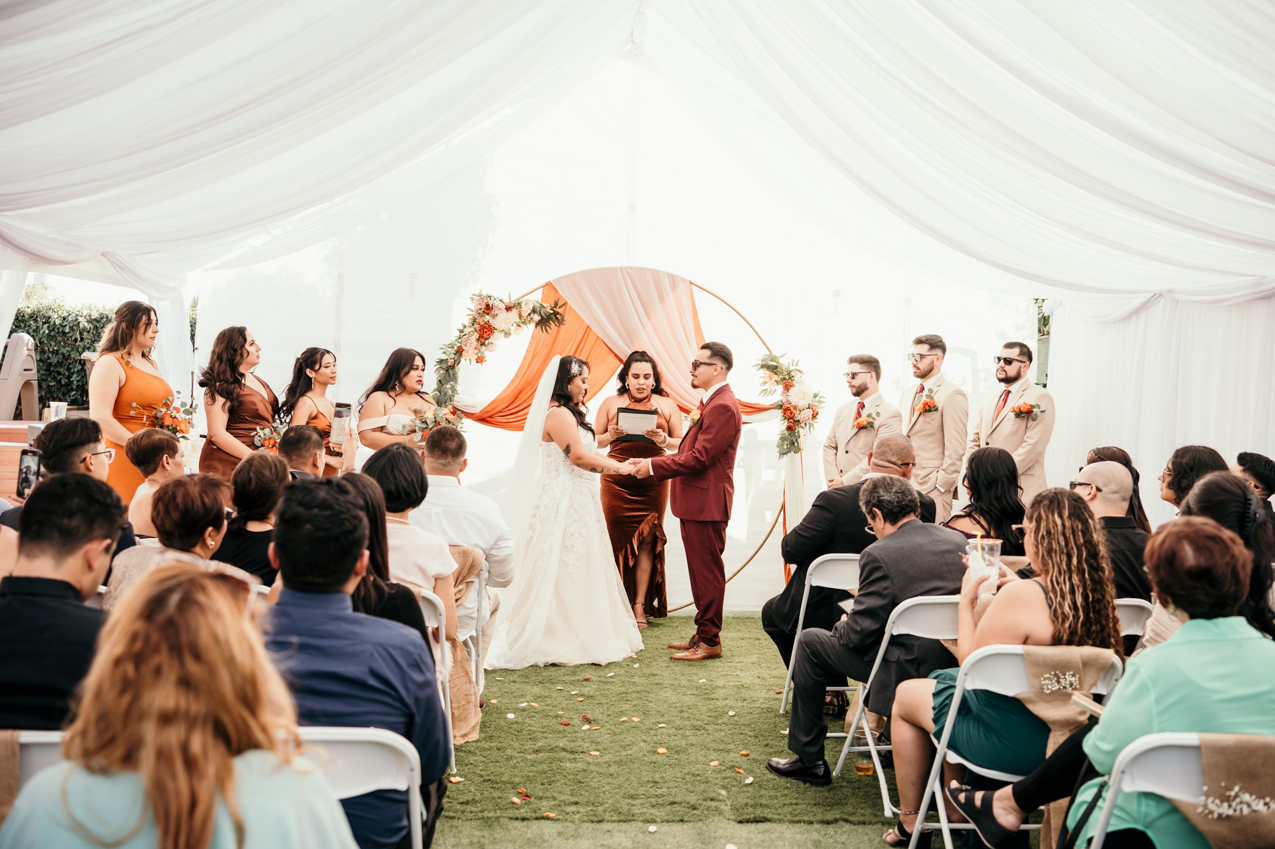 A wedding ceremony takes place under an elegant white tent with guests seated in folding chairs on a grassy area.