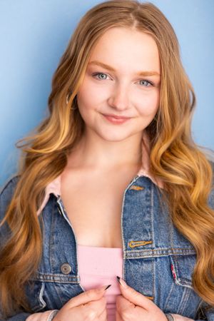 Young woman with long wavy auburn hair wearing a denim jacket over pink top, smiling against blue background.