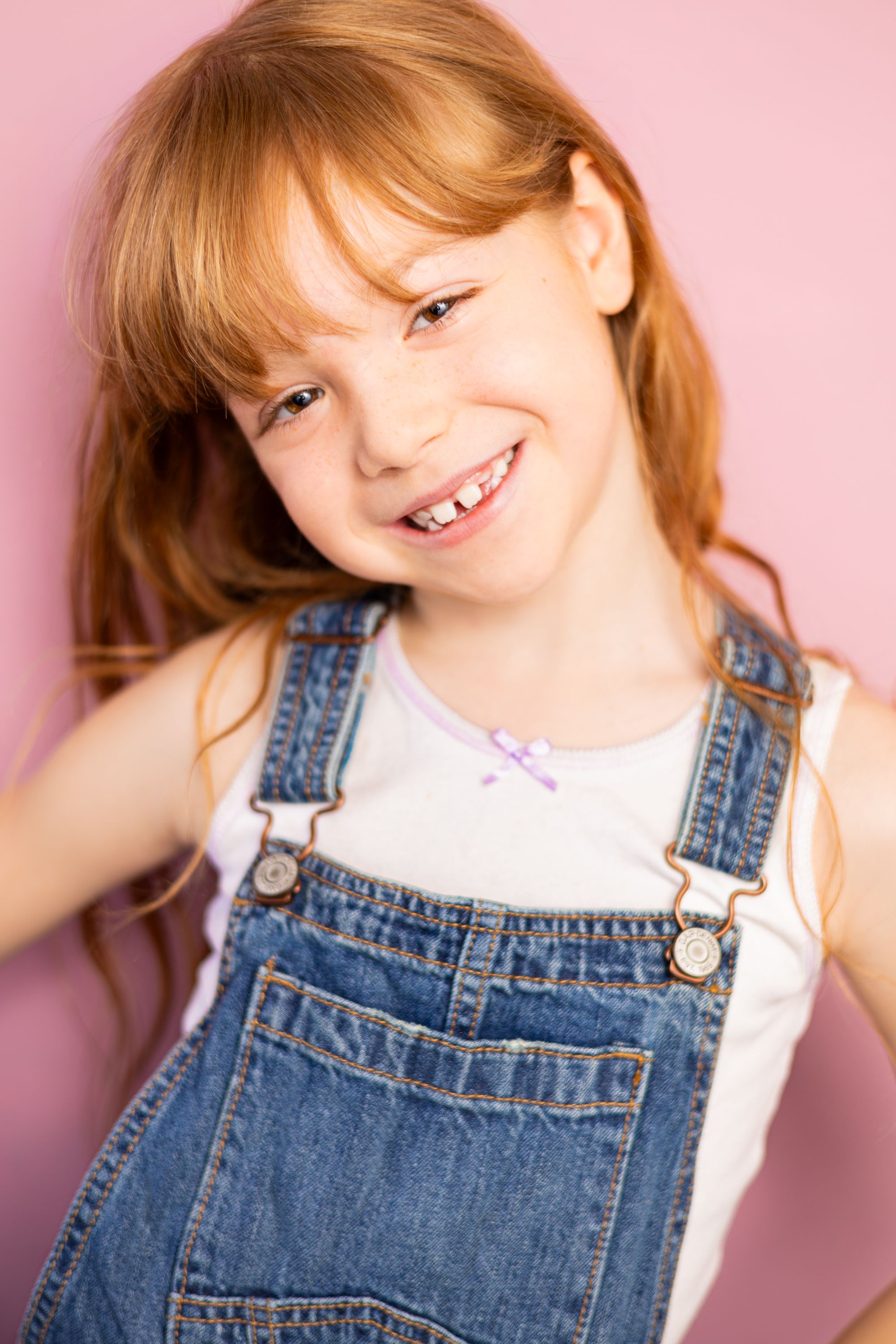 Smiling redhead girl in denim overalls and white shirt posing against a pink background.