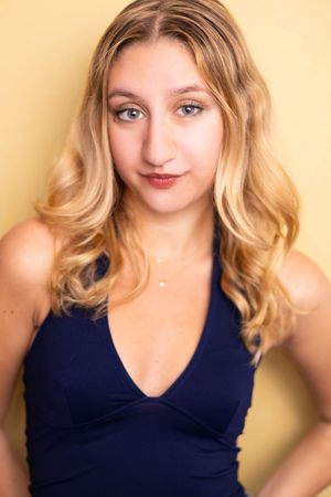 Young blonde woman with wavy hair wearing a navy blue halter top poses against a yellow background.