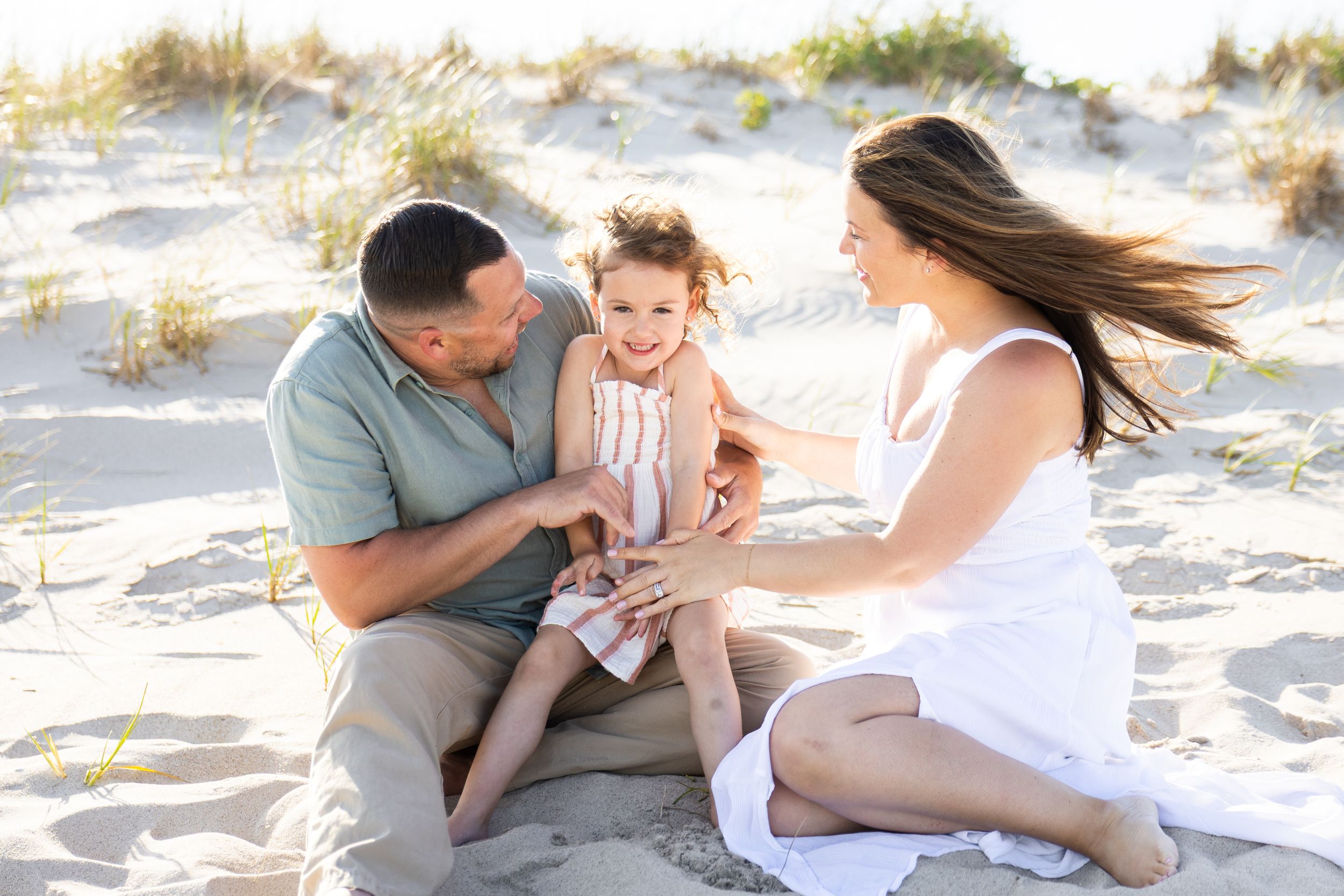 A happy family of three sits on sandy beach dunes, parents smiling at their toddler daughter on a sunny day.