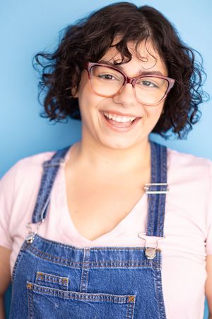 Smiling young woman with curly hair, glasses, and denim overalls against a blue background.