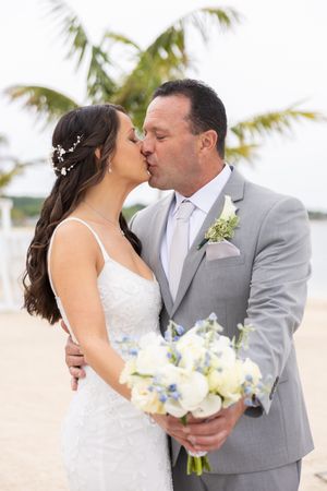 Bride and groom share a kiss on a beach, she holds white and blue bouquet, he wears a gray suit with boutonniere.