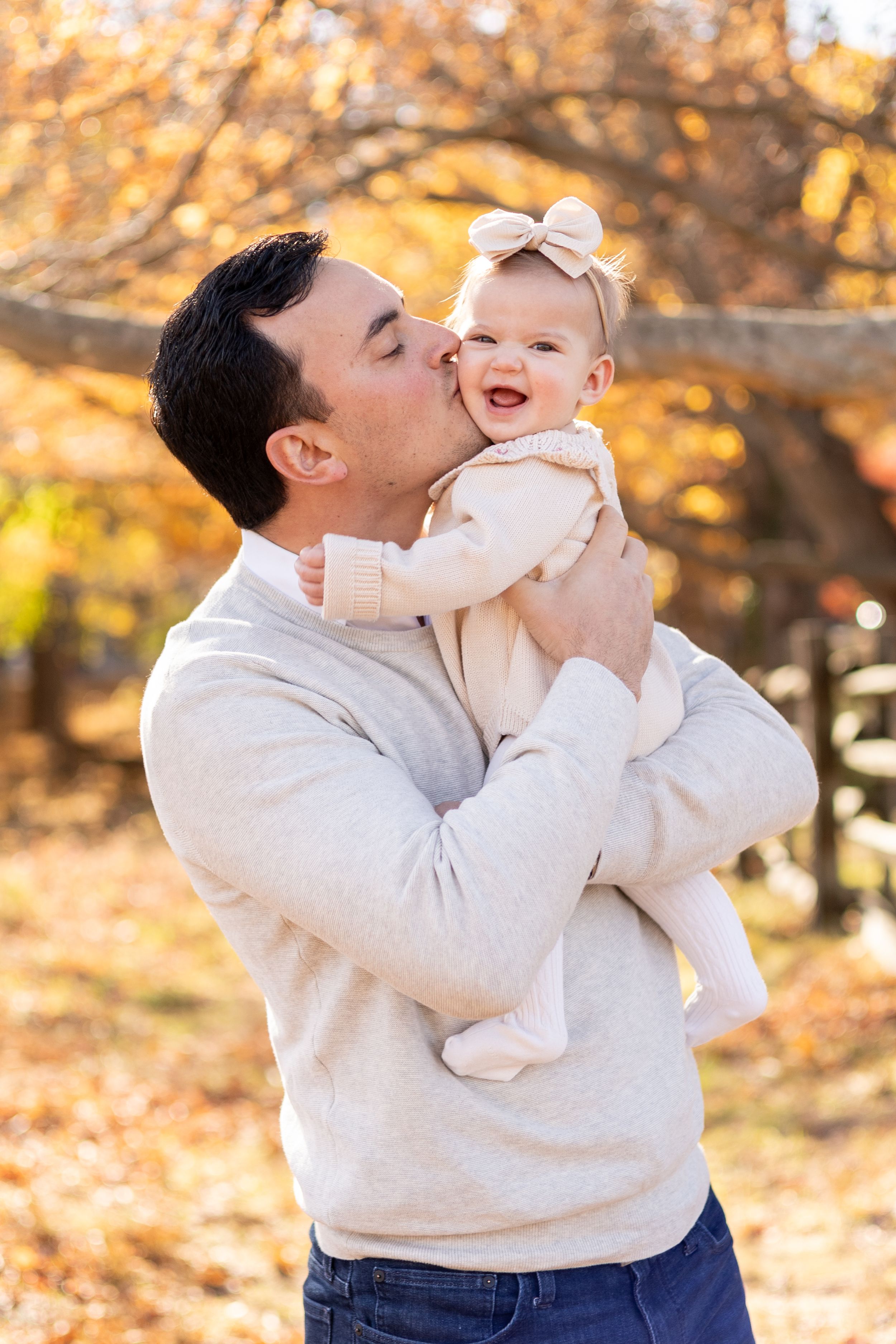 Father kissing his smiling baby girl outdoors among colorful autumn foliage.