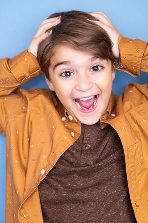 Excited young boy in an orange jacket with hands on head laughing against a blue background.