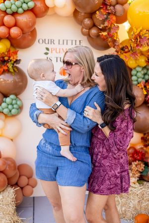 Two women and a baby celebrate a fall-themed first birthday party in front of a colorful balloon arch.