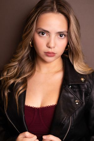 Young woman with long highlighted wavy hair in black leather jacket and burgundy camisole on dark background.