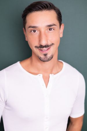 Young man with goatee wearing white henley shirt poses against dark teal background in professional headshot.