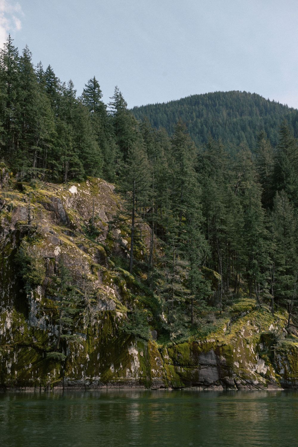 Rocky cliff face covered in evergreen trees along pristine mountain lake shoreline.