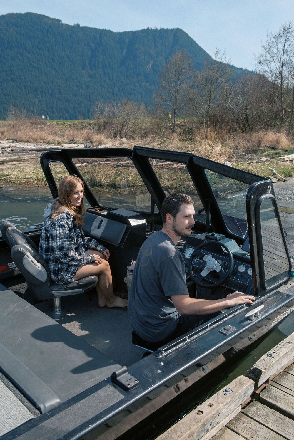 People relaxing in black boat on wooden dock with mountain backdrop.