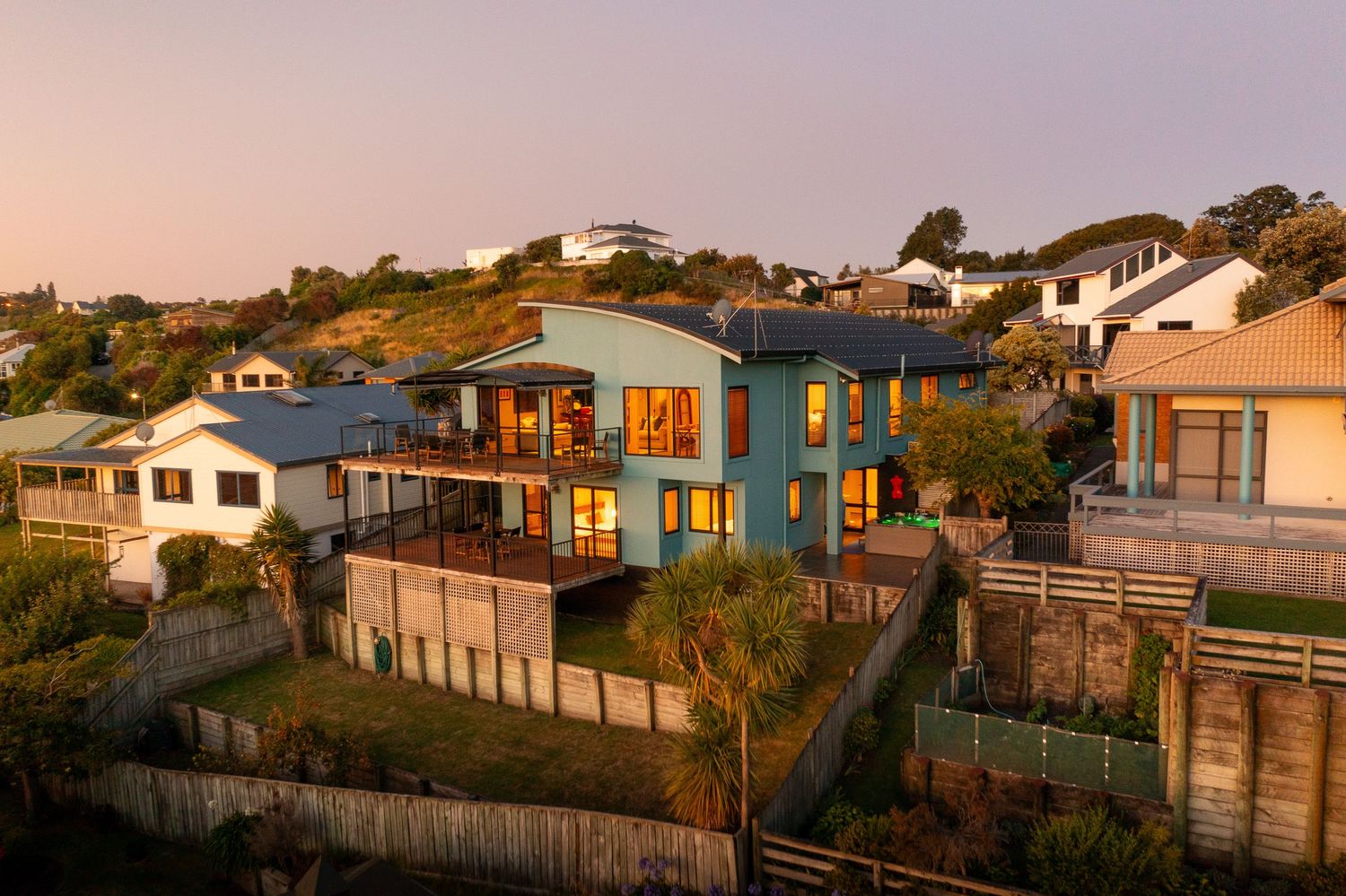 Modern hillside home with blue exterior glows warmly at sunset in a coastal residential neighborhood.