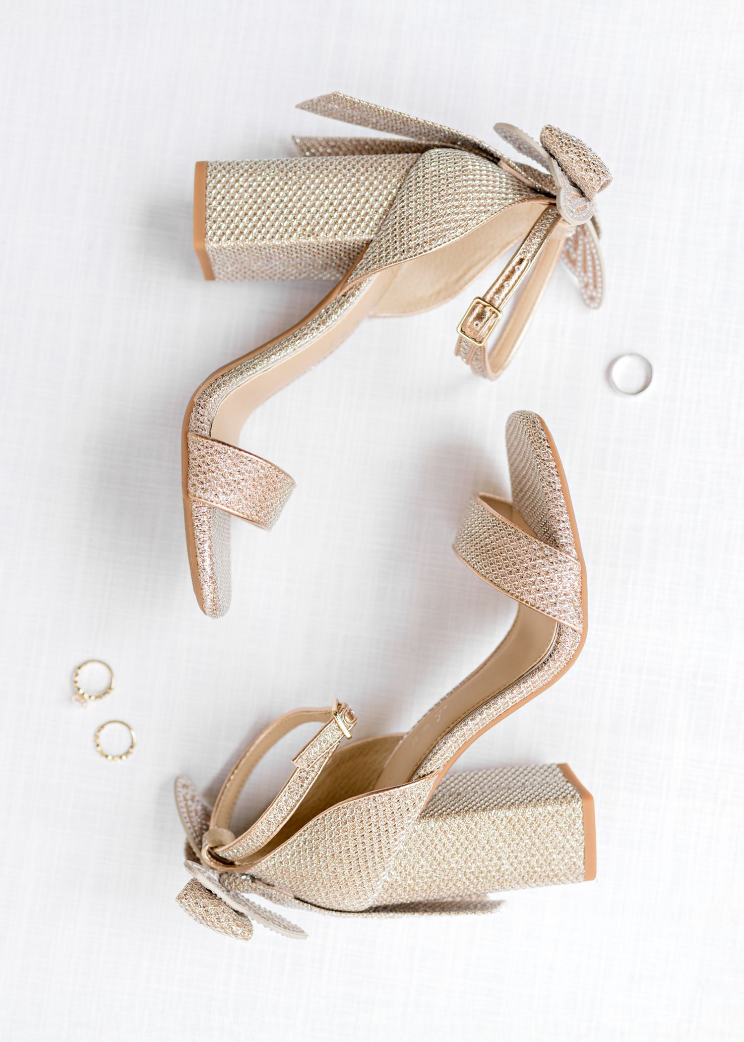 Elegant nude block heel sandals with ankle straps and bows displayed with delicate gold rings on white background.