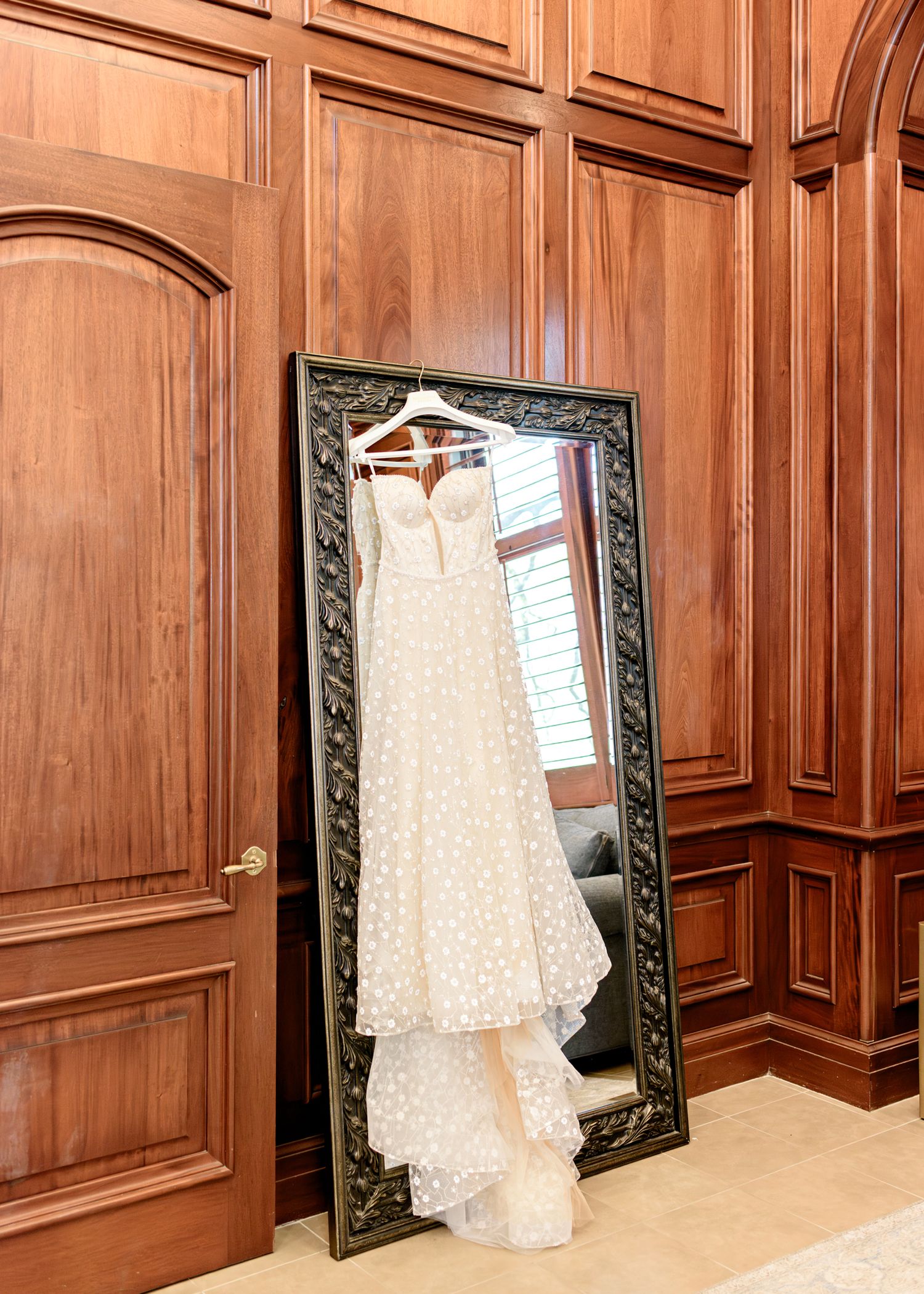 An elegant white lace wedding dress hangs on display against an ornate mirror with detailed wooden wall paneling.