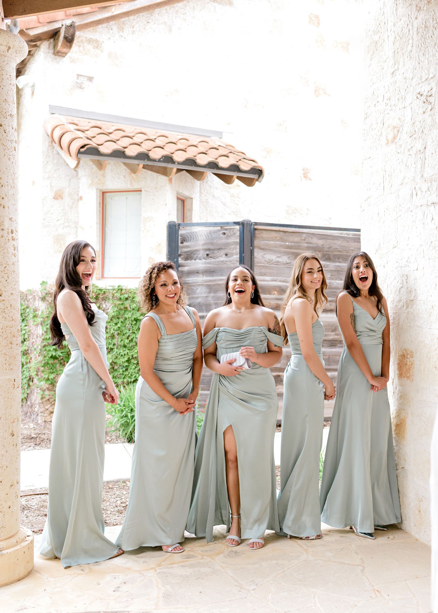 Five bridesmaids wearing elegant sage green dresses laugh together under a Spanish-style archway.