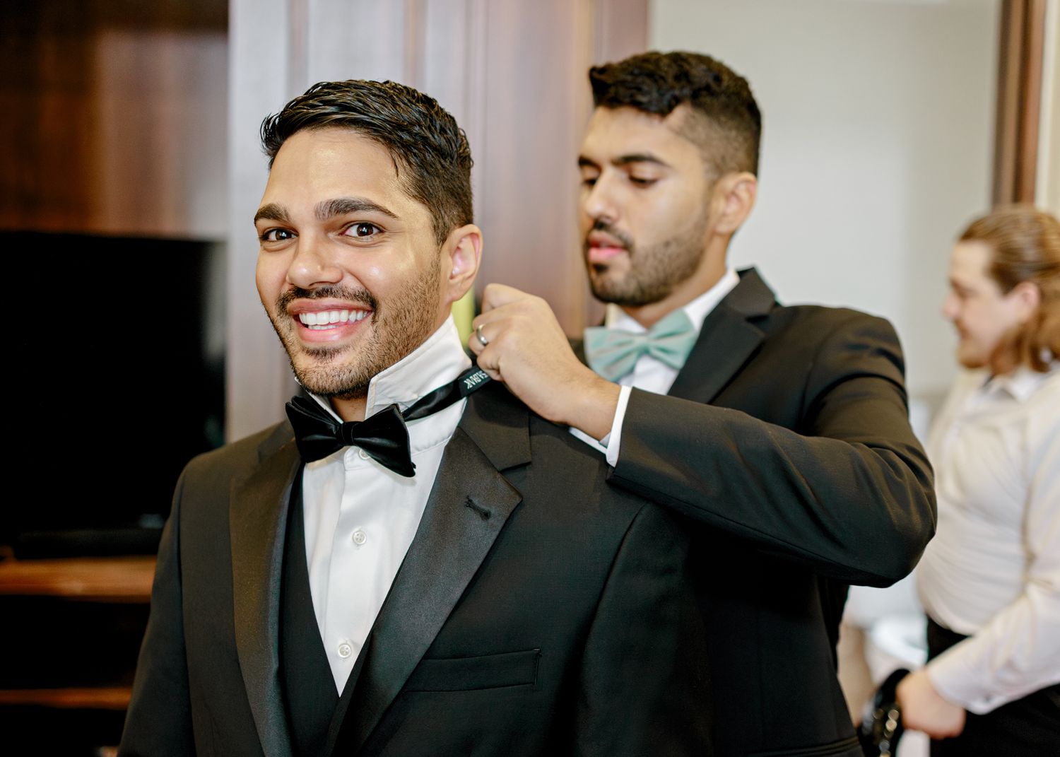 A groom in a black tuxedo and bowtie smiles while someone helps adjust his collar during wedding preparations.